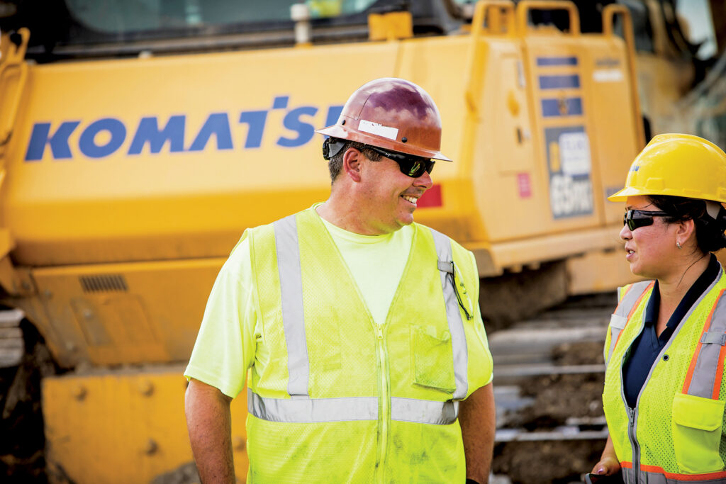 A man and a woman working on a construction job site in the heat in front of a Komatsu D65PXi