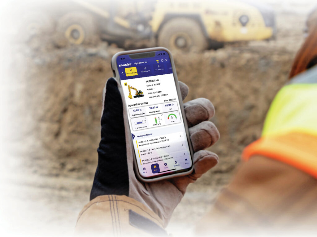 A gloved construction worker holding a phone with the My Komatsu mobile app interface on it