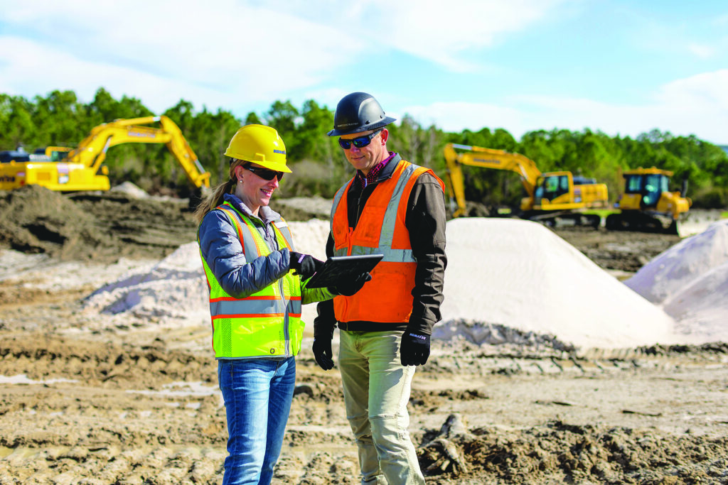 Smart Construction solutions: A man and a women in hard hats and vests standing in front of Komatsu excavators looking at information on an ipad