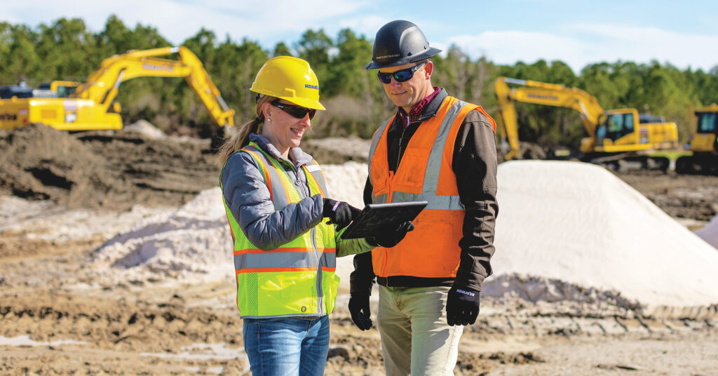 Smart Construction solutions: A man and a women in hard hats and vests standing in front of Komatsu excavators looking at information on an ipad