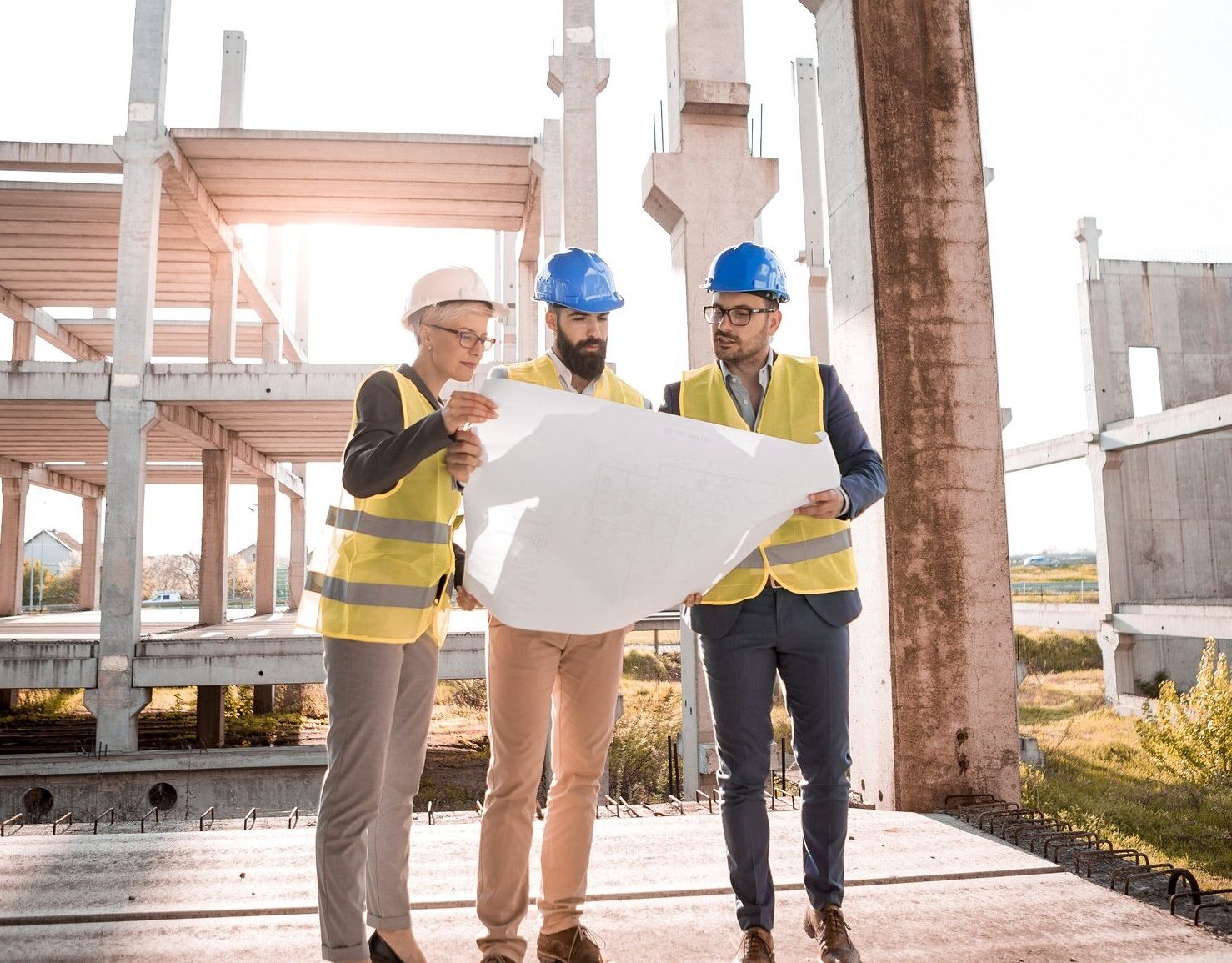 3 men in hard hats and vests stand in front of a building that is under construction all looking at a building plan on paper