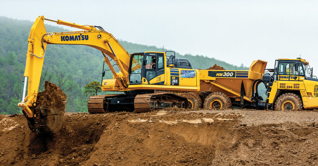 A Komatsu Hybrid HB365LC-3 Excavator moving dirt next to a Komatsu HM300 Truck