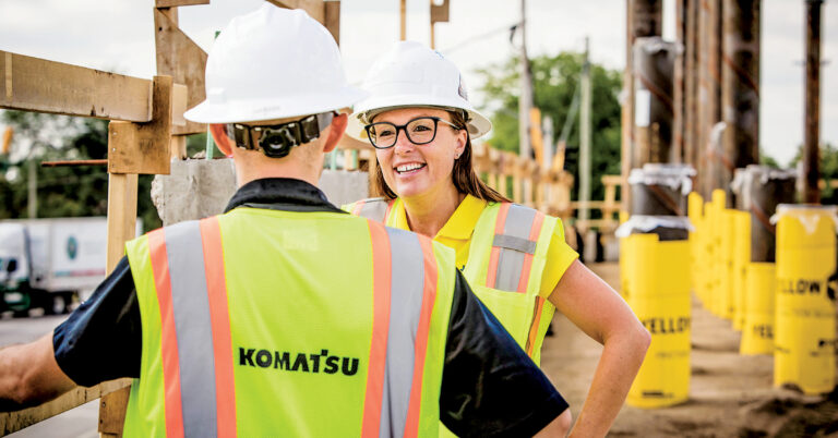 Construction Site Injuries | Two construction workers in Komatsu safety vests talk at a job site