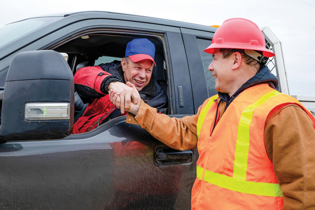two men, one in a truck, and the other outside the truck and wearing a hard hat and safety vest shaking hands through the truck window