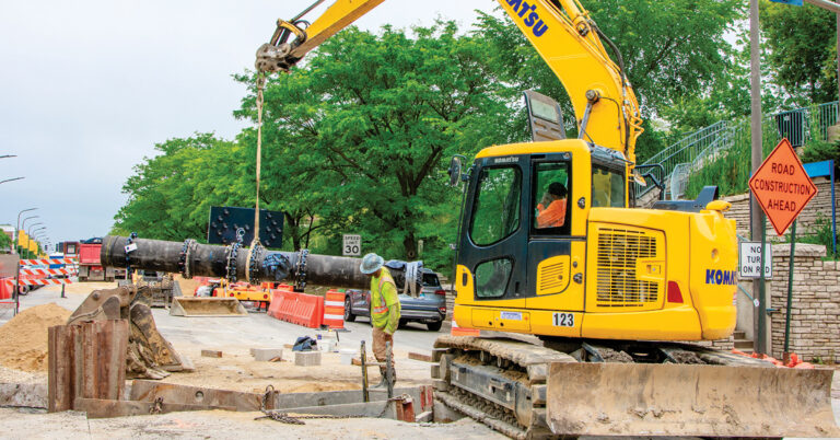The benefit of Infrastructure investment is shown with a Komatsu excavator moving pipe within a roadwork zone